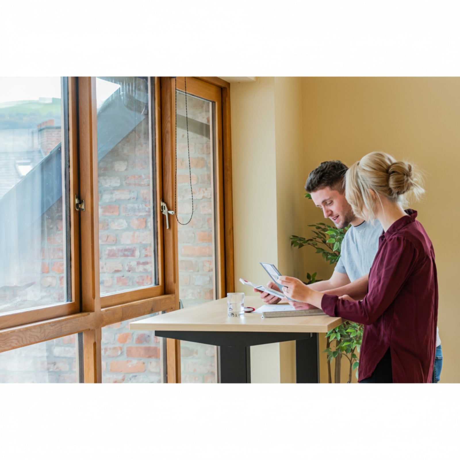 Man and woman collaborating at Quiet Motor Standing Desk by window reviewing paperwork