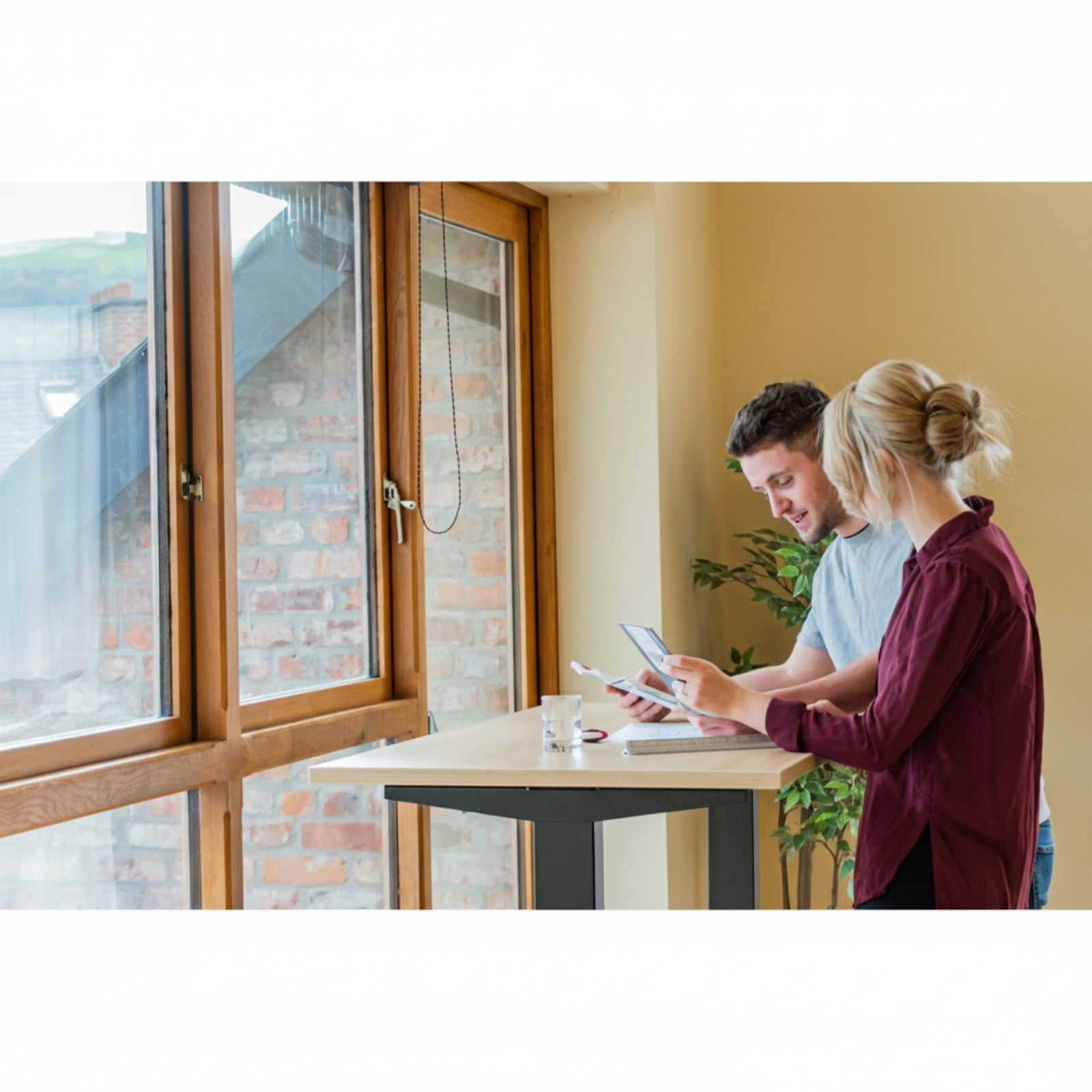Man and woman collaborating at Quiet Motor Standing Desk by window reviewing paperwork