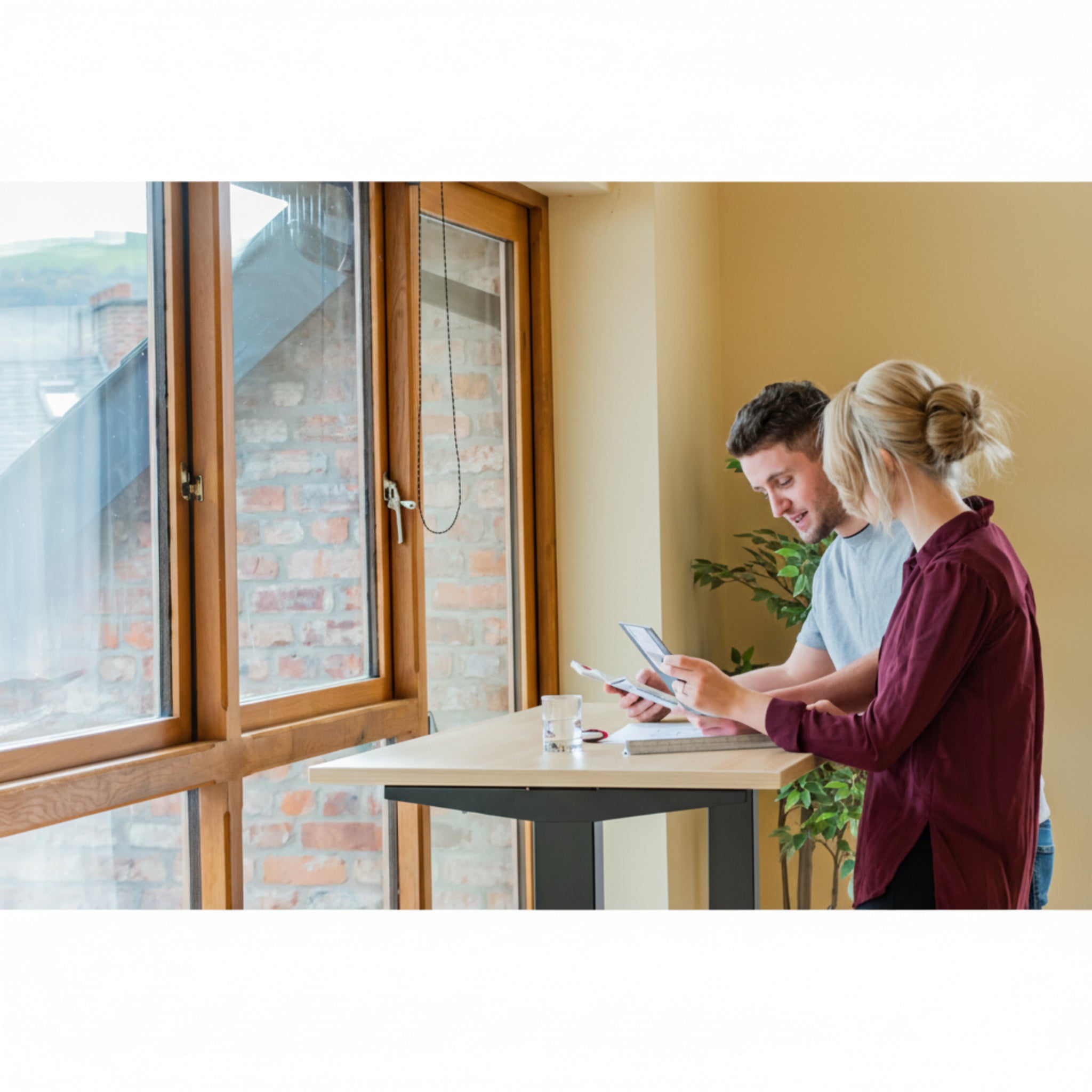 Man and woman collaborating at Quiet Motor Standing Desk by window reviewing paperwork