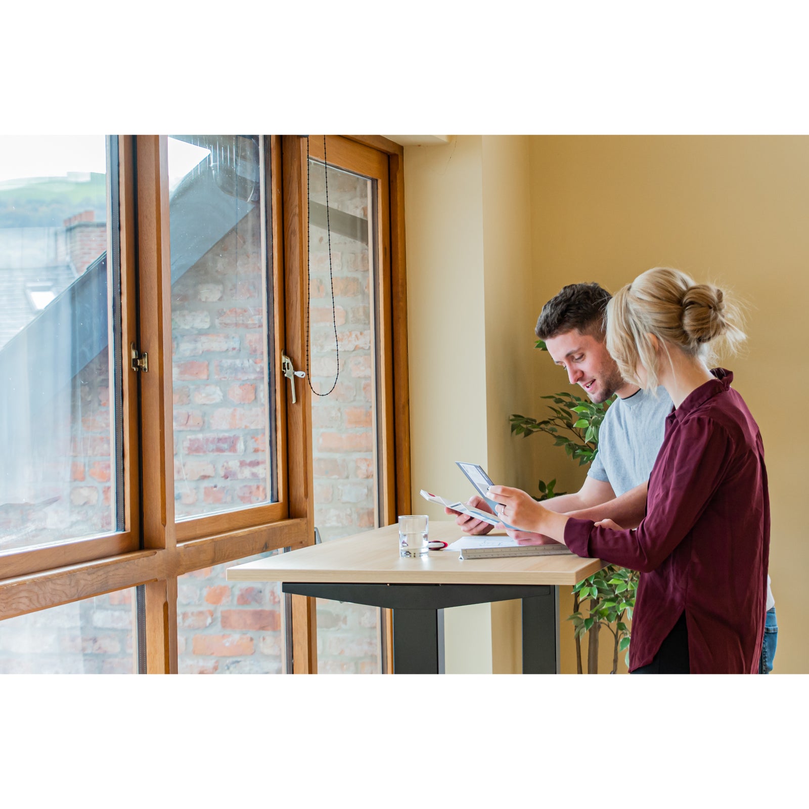 Man and woman collaborating at Quiet Motor Standing Desk by window reviewing paperwork