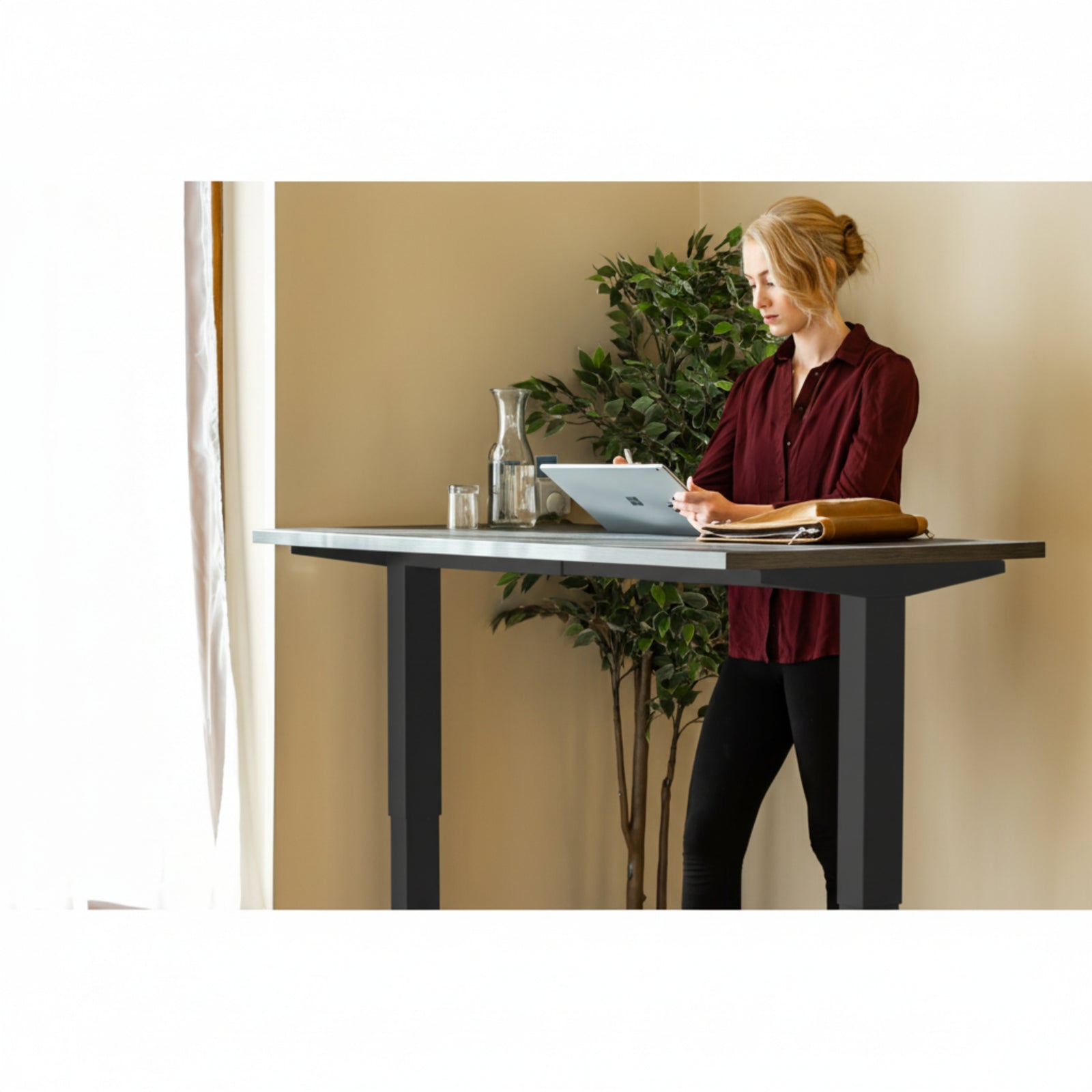 Woman using tablet while standing at Quiet Motor Standing Desk with a tall plant behind her