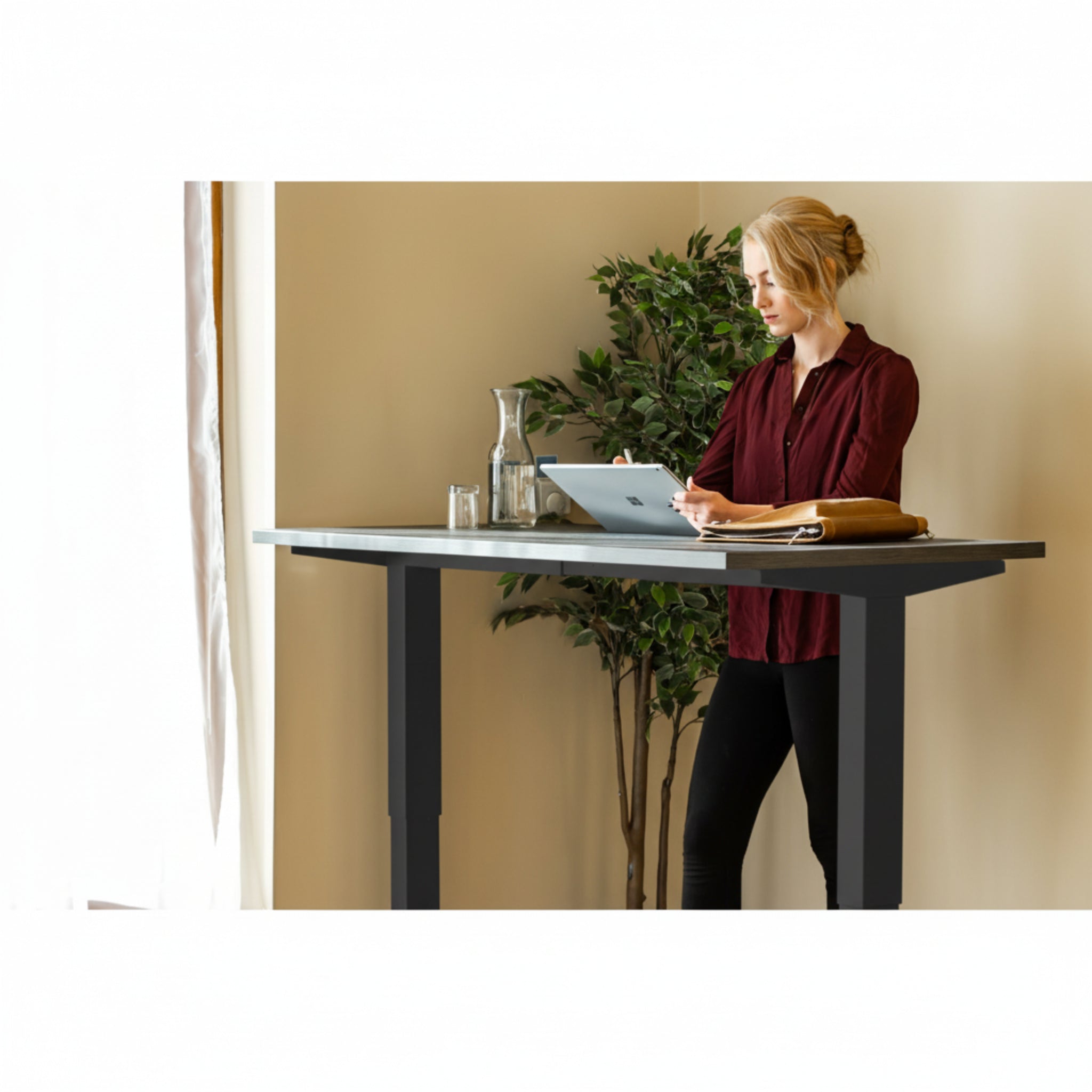 Woman using tablet while standing at Quiet Motor Standing Desk with a tall plant behind her