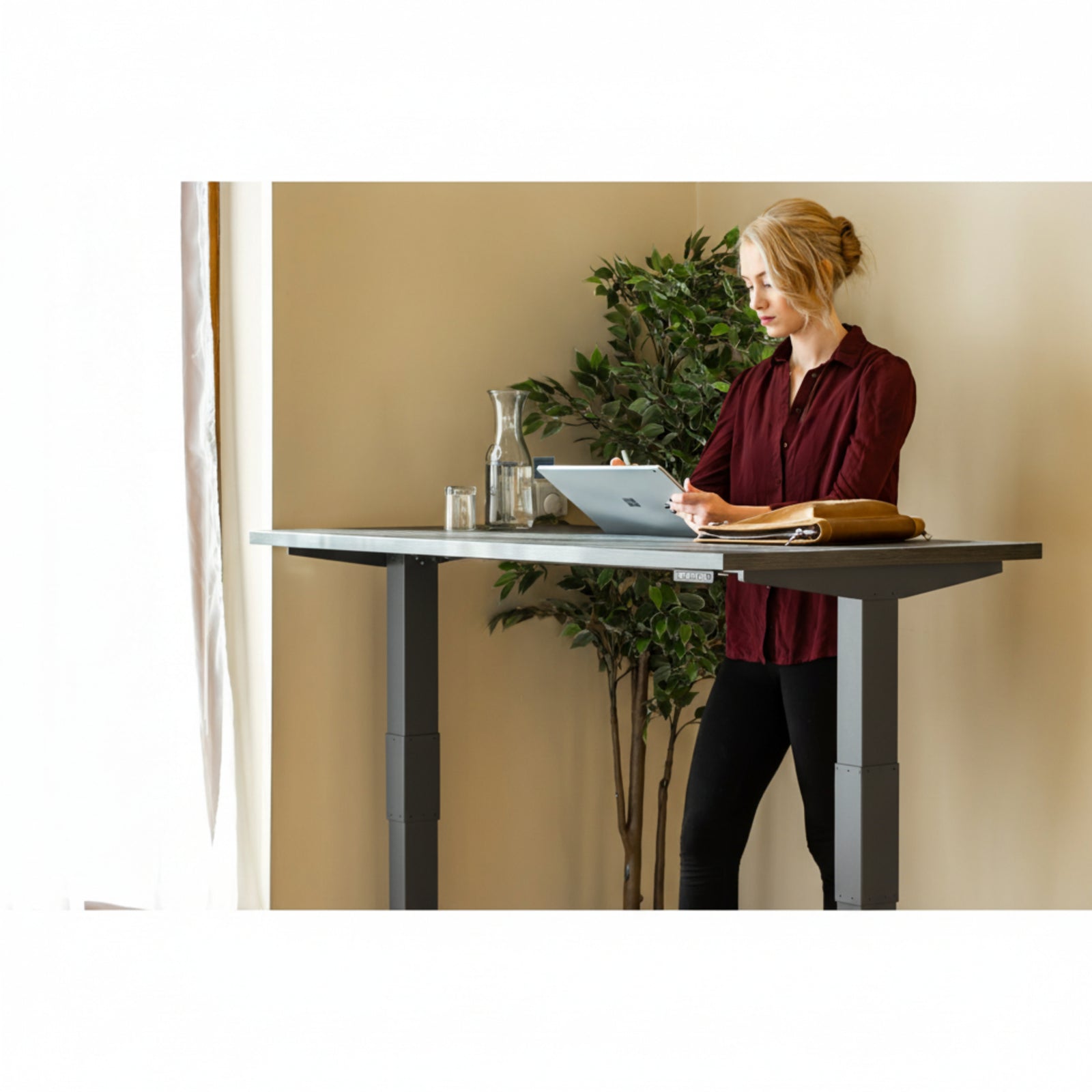 Woman using tablet while standing at Quiet Motor Standing Desk with a tall plant behind her