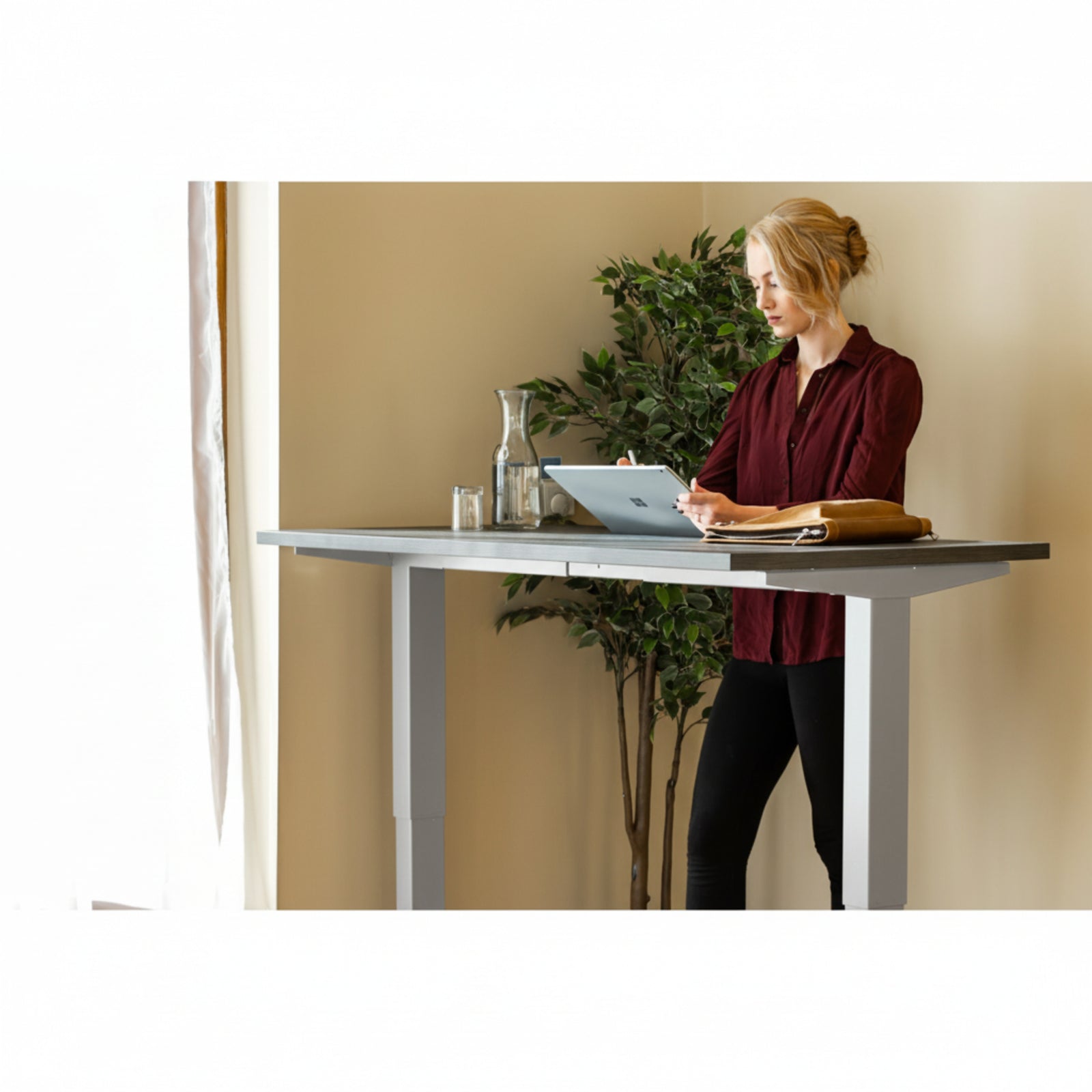 Woman using tablet while standing at Quiet Motor Standing Desk with a tall plant behind her