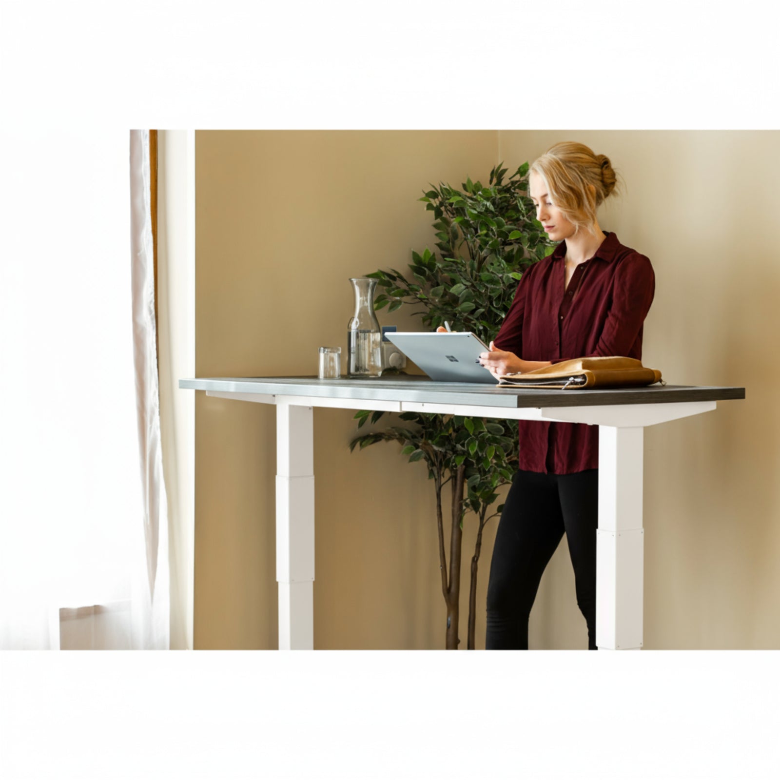 Woman using tablet while standing at Quiet Motor Standing Desk with a tall plant behind her