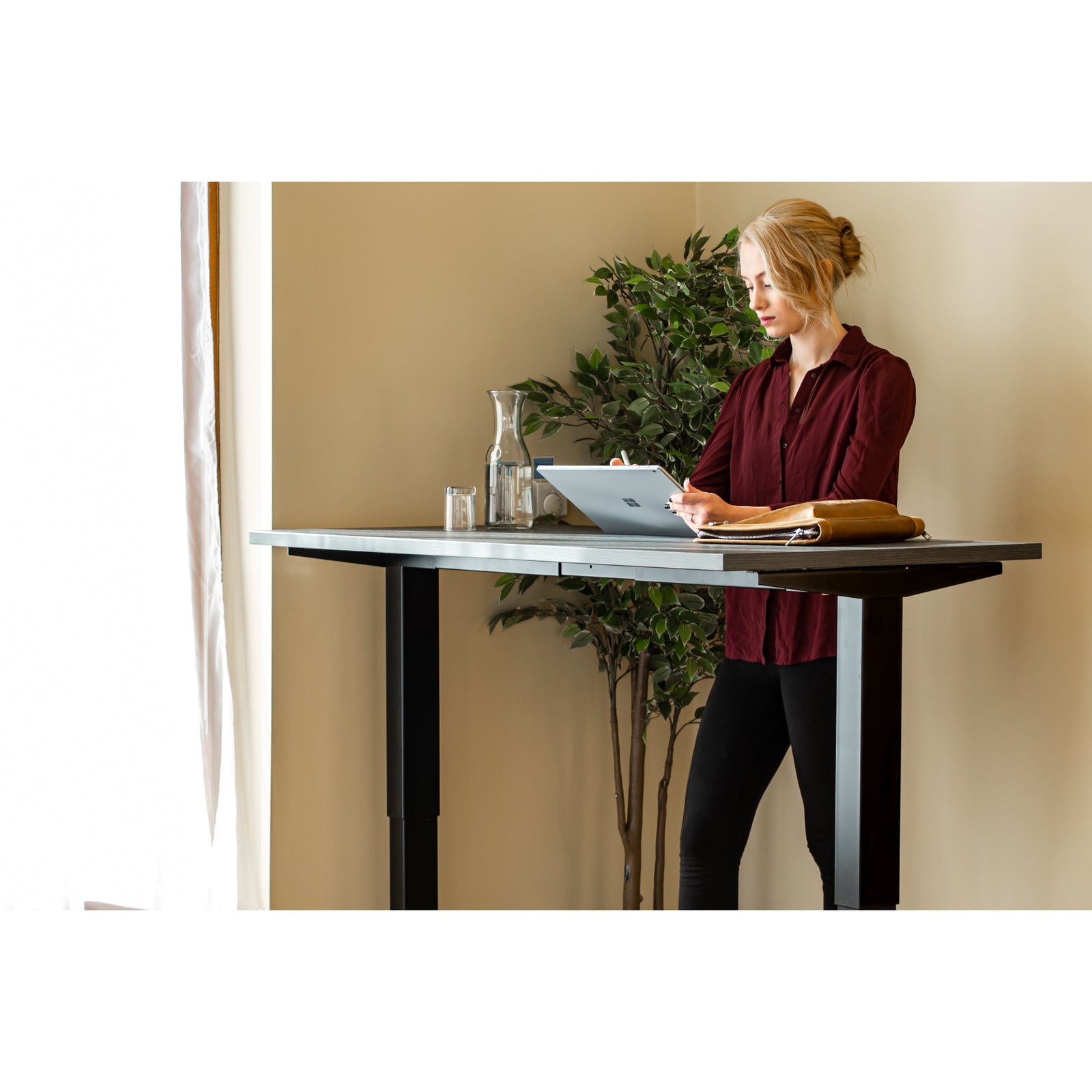 Woman using tablet while standing at Quiet Motor Standing Desk with a tall plant behind her