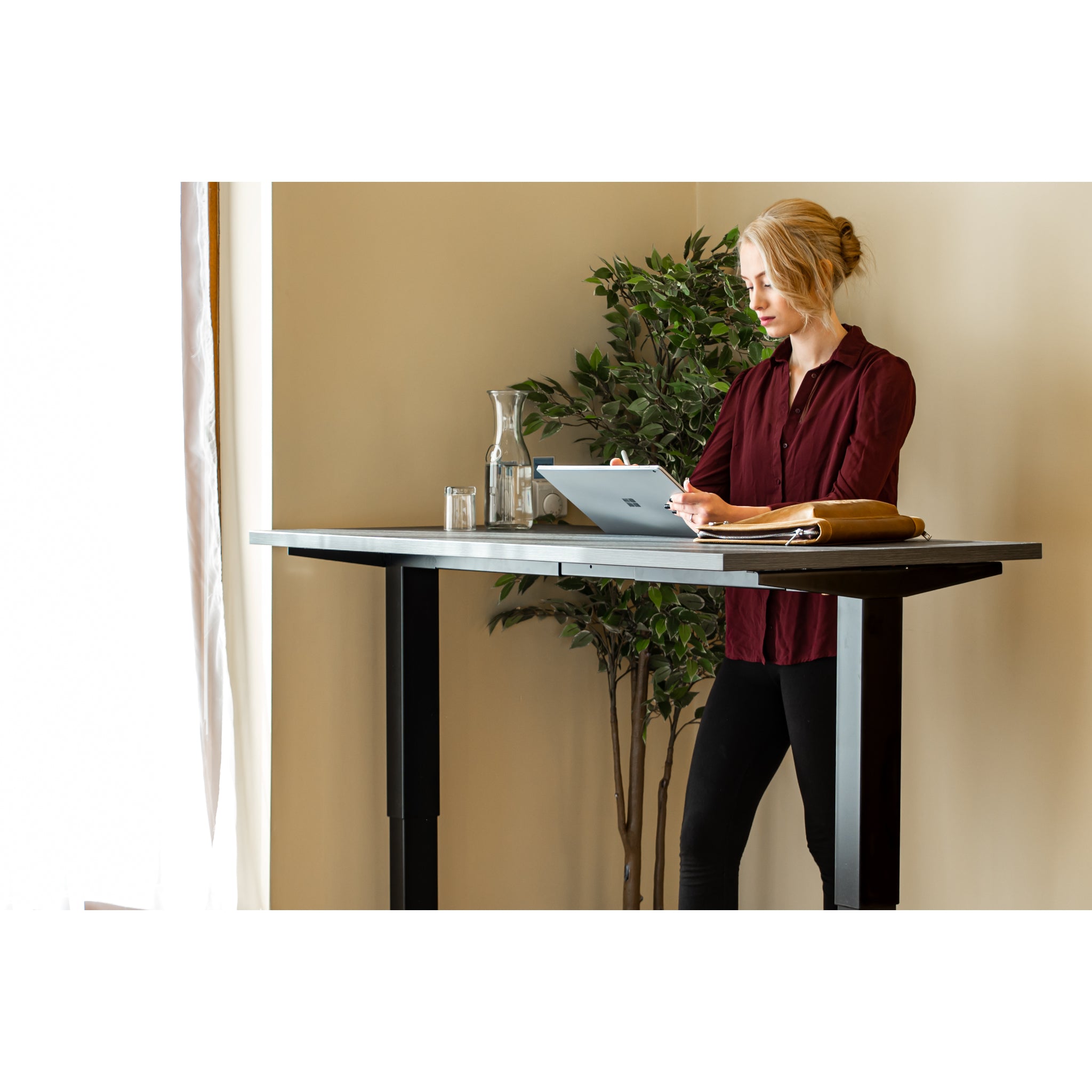 Woman using tablet while standing at Quiet Motor Standing Desk with a tall plant behind her