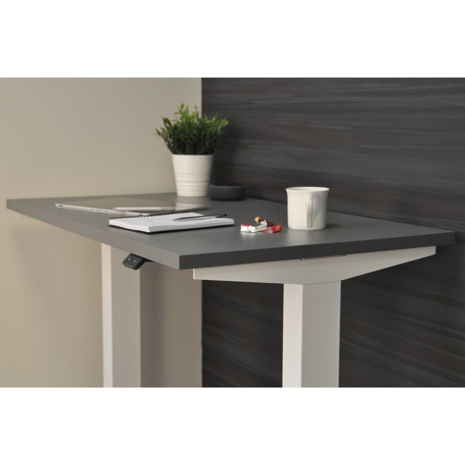 UrbanErgo white frame standing desk with grey desktop, shown in a compact modern home office with a potted plant and notepad