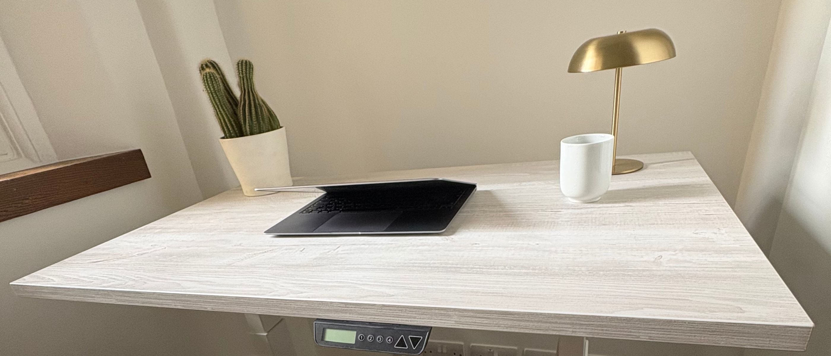 UrbanErgo Core Standing Desk in a compact home office with white frame, laptop, brass lamp and cactus