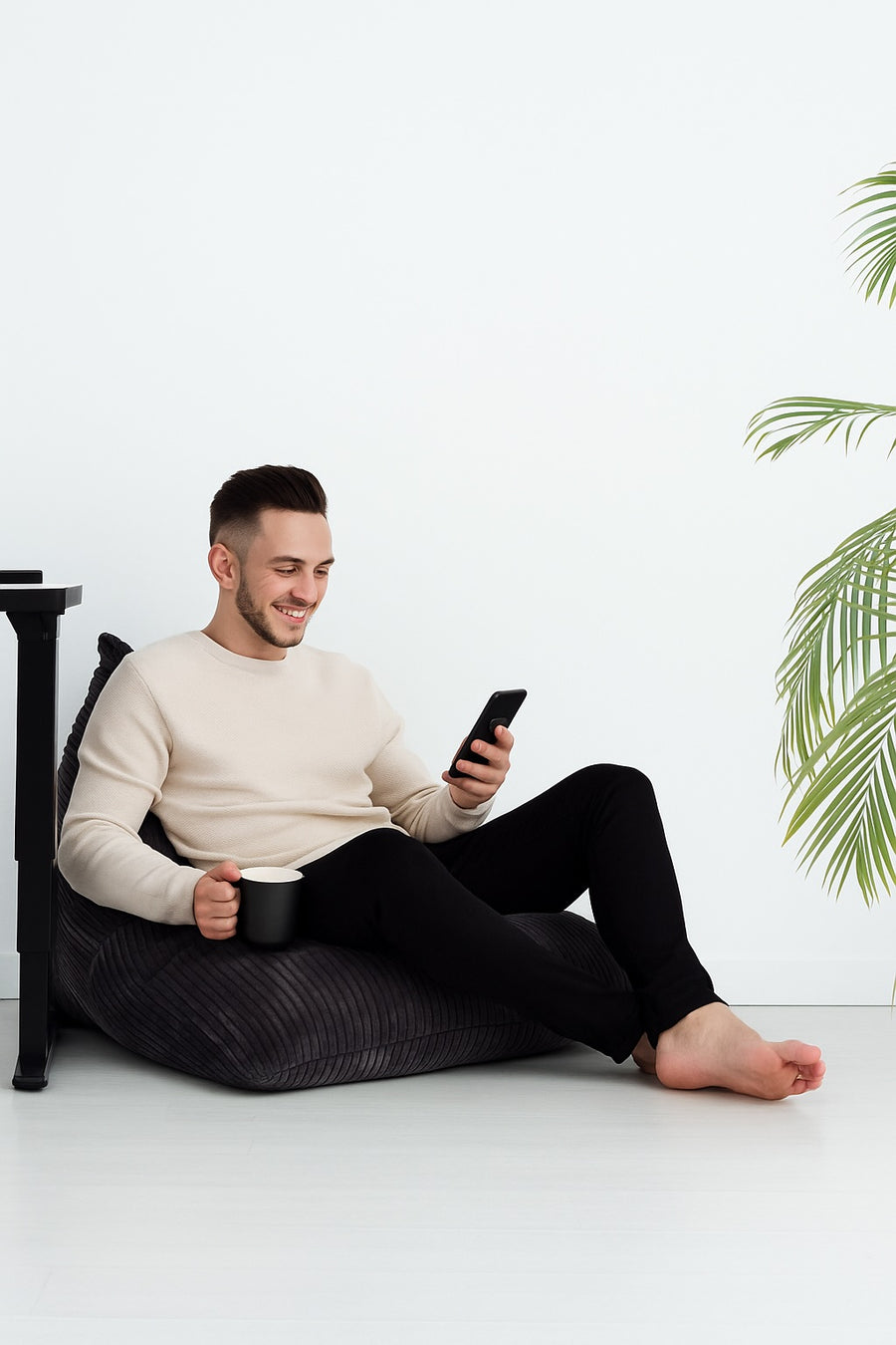 A man relaxing on a beanbag with a coffee mug and smartphone next to a black standing desk and indoor plants in a minimalist home office