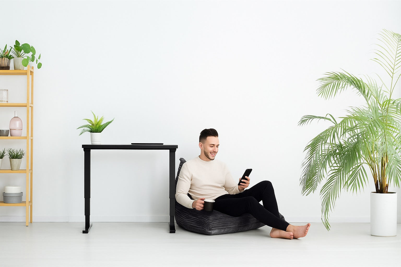 A man relaxing on a beanbag with a coffee mug and smartphone next to a black standing desk and indoor plants in a minimalist home office